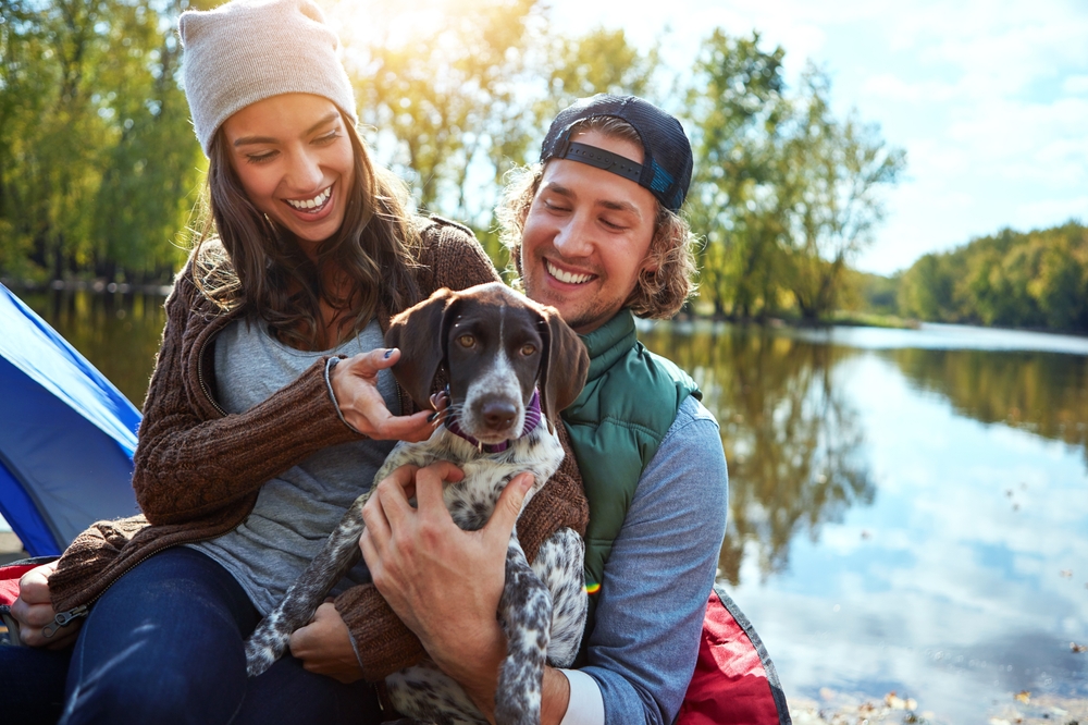 Couple with dog enjoying nature by the water – River Grove Animal Hospital Smiling couple sitting by a peaceful riverbank with a blue tent behind them, holding a spotted dog between them as sunlight filters through the trees – River Grove Animal Hospital